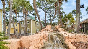 A natural rock formation with a small waterfall, surrounded by palm trees and a building with a green roof. - Family-friendly Pensacola Home < 1 Mi to Beach! (Pensacola)