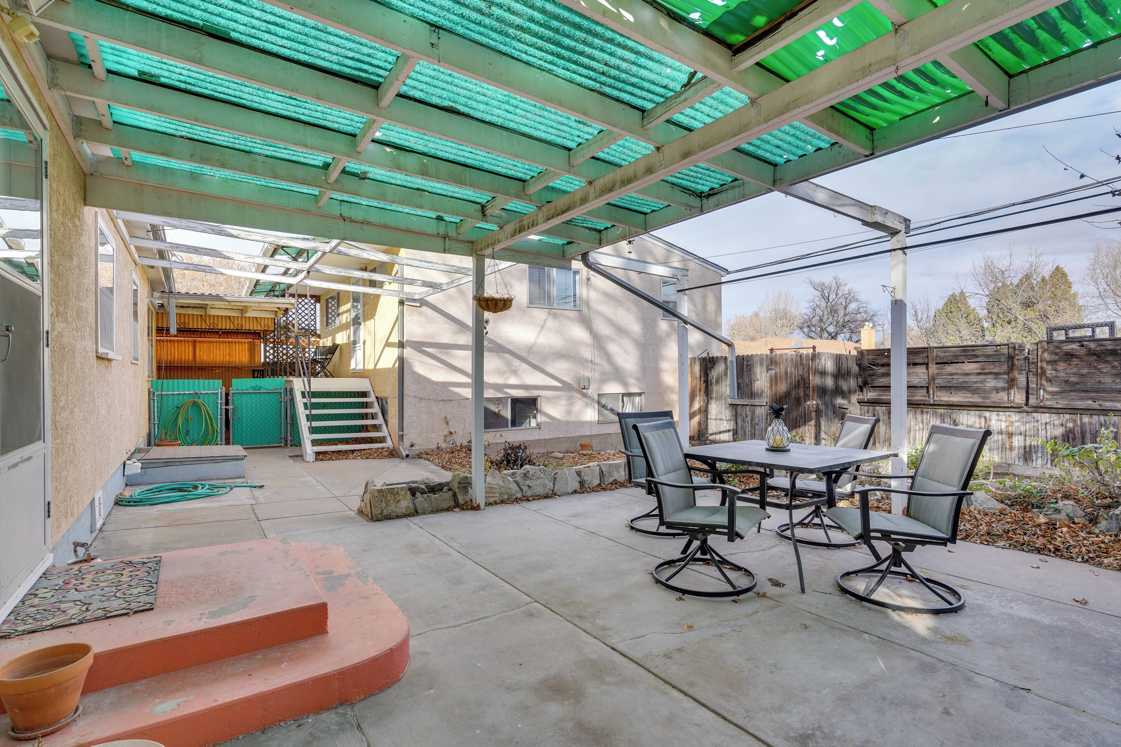 A covered patio area with a table and chairs, a hanging basket, and a wooden fence in the background.