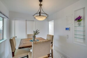 Zona pranzo con tavolo in legno, sedie imbottite color beige, lampadario, pianta in vaso e ripiano in vetro con oggetti decorativi.
