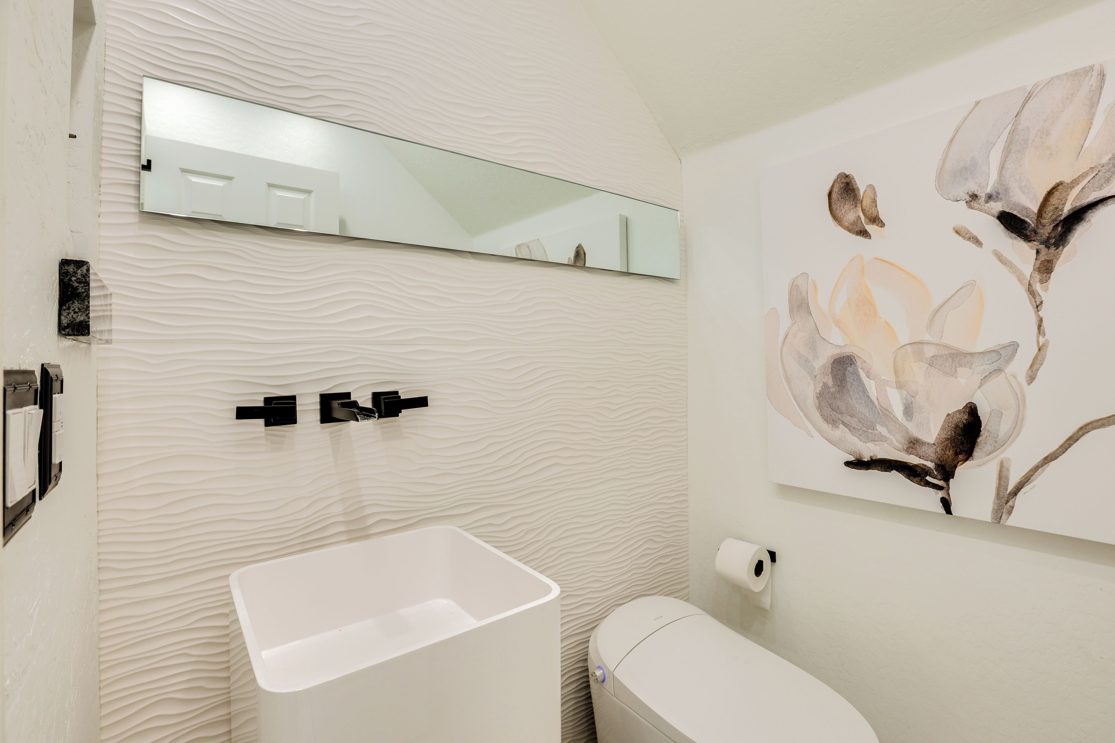 A modern bathroom with a textured wall, a rectangular sink, a toilet, a mirror, and a floral artwork.