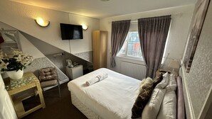 A hotel room with a bed, a desk with a chair, a TV mounted on the wall, and a window with curtains. - Ocean Road Rooms (South Shields)