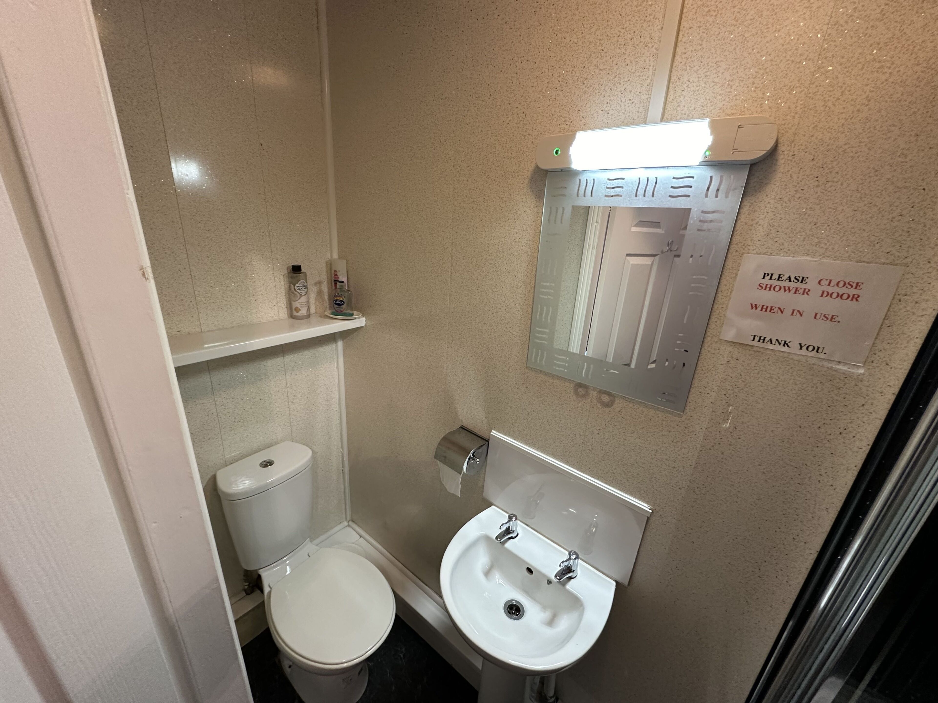 Un baño pequeño con inodoro, lavamanos y espejo. Hay un cartel que solicita cerrar la puerta de la ducha al usarla.