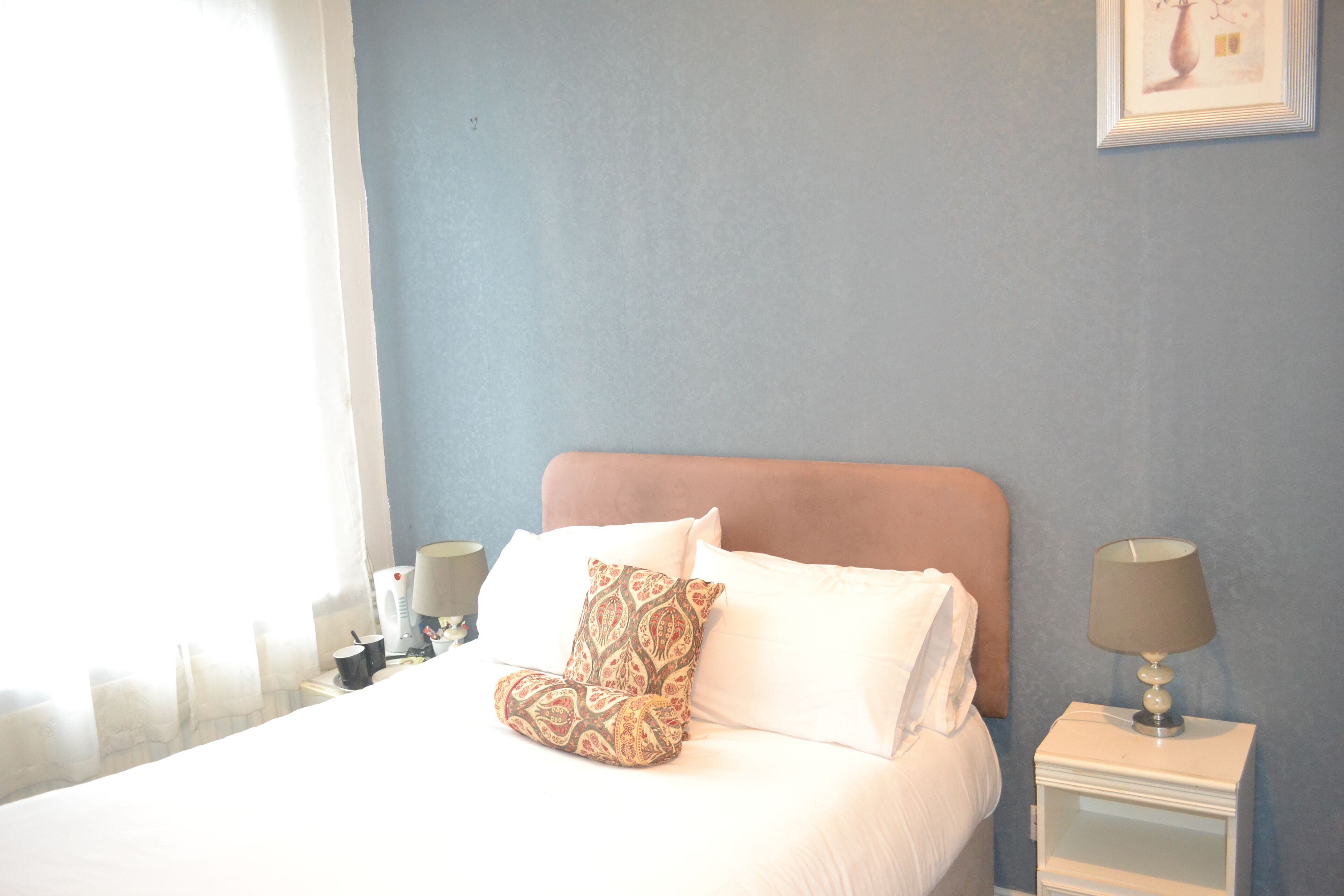 A neatly made bed with white linens and a patterned pillow, a bedside lamp, and a framed picture on the wall.