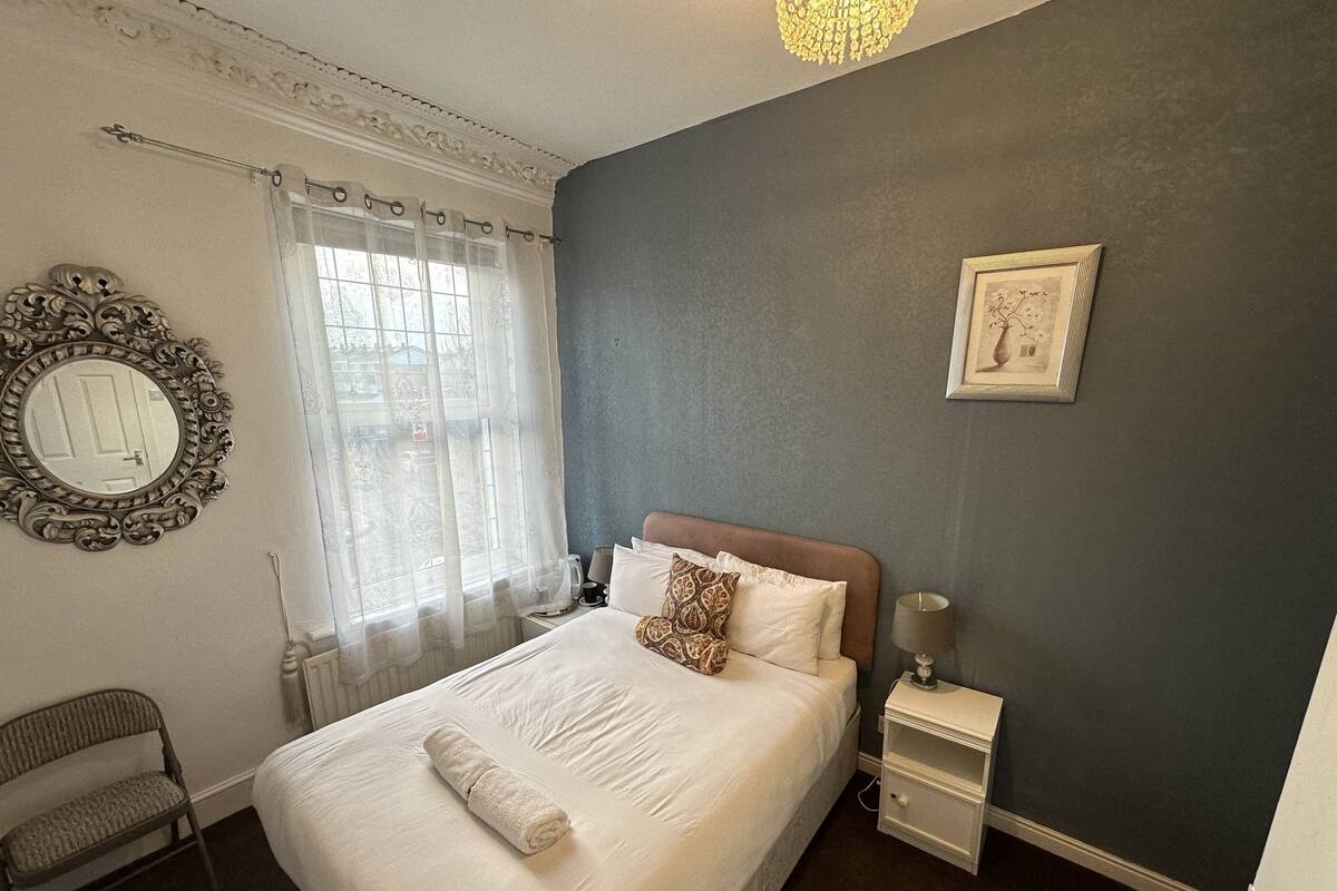 A neatly made bed with a rolled towel, a bedside table with a lamp, a chair, and a framed picture on the wall.