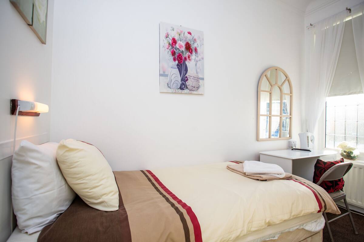 A neatly arranged hotel room with a bed, a desk, a chair, a window with curtains, and a wall-mounted artwork.
