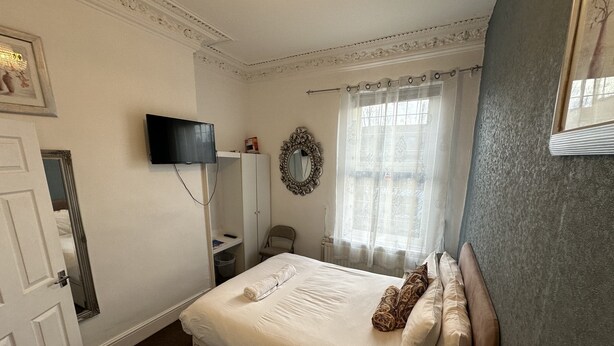A bedroom with a bed, a television, a wardrobe, a chair, and a window with curtains. - Ocean Road Rooms (South Shields)