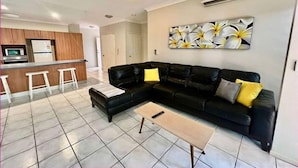 Een moderne woonkamer met een zwarte leren bank, een houten salontafel, en een keuken met een witte koelkast en houten kastjes.