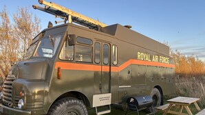 Vanhanmallinen, armeijatyylinen ajoneuvo, jonka kylkeen on maalattu teksti ”ROYAL AIR FORCE”, on pysäköity nurmikolle. Lähellä on piknikpöytä ja grilli.