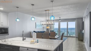 A modern kitchen with a large island, dark cabinetry, and a dishwasher. There is a dining area with a view of the ocean and a balcony with outdoor seating. - Luxury Beachfront Bliss – Blu’s Only 2BR Condo! (Fort Walton Beach)