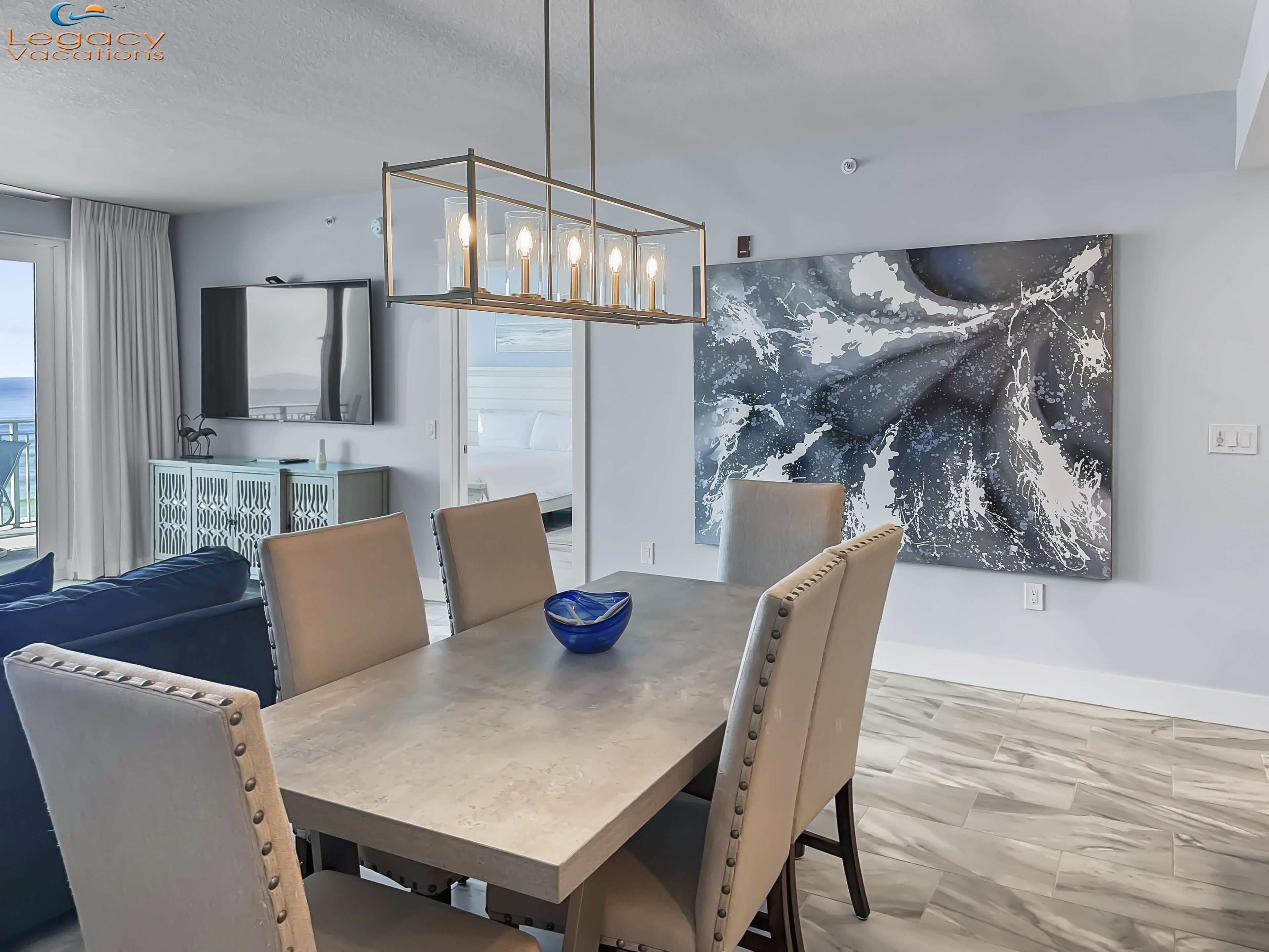 A modern dining area with a wooden table, beige upholstered chairs, and a blue bowl. A large abstract painting adorns the wall, and a television is mounted above the table.