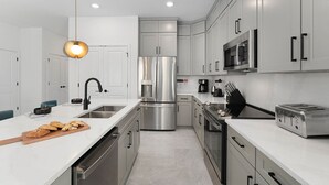 A modern kitchen with stainless steel appliances, a white countertop, and a hanging pendant light. - Villa Bellisima | Huge I-Drive villa w PVT pool, grand BRs, luxe LR, near parks (Orlando)