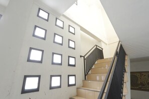 Una scala con ringhiera nera e gradini color beige, che conduce a un pianerottolo con parete bianca e diverse finestre quadrate.