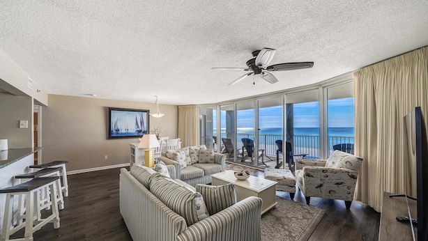 A spacious living room with a large sofa, a coffee table, and a TV. There is a balcony with a view of the ocean. - FREE Beach Service, Golf &Perks! Long Beach Tower 3-303, Hot Tub, Gym & Pools (Panama City Beach)