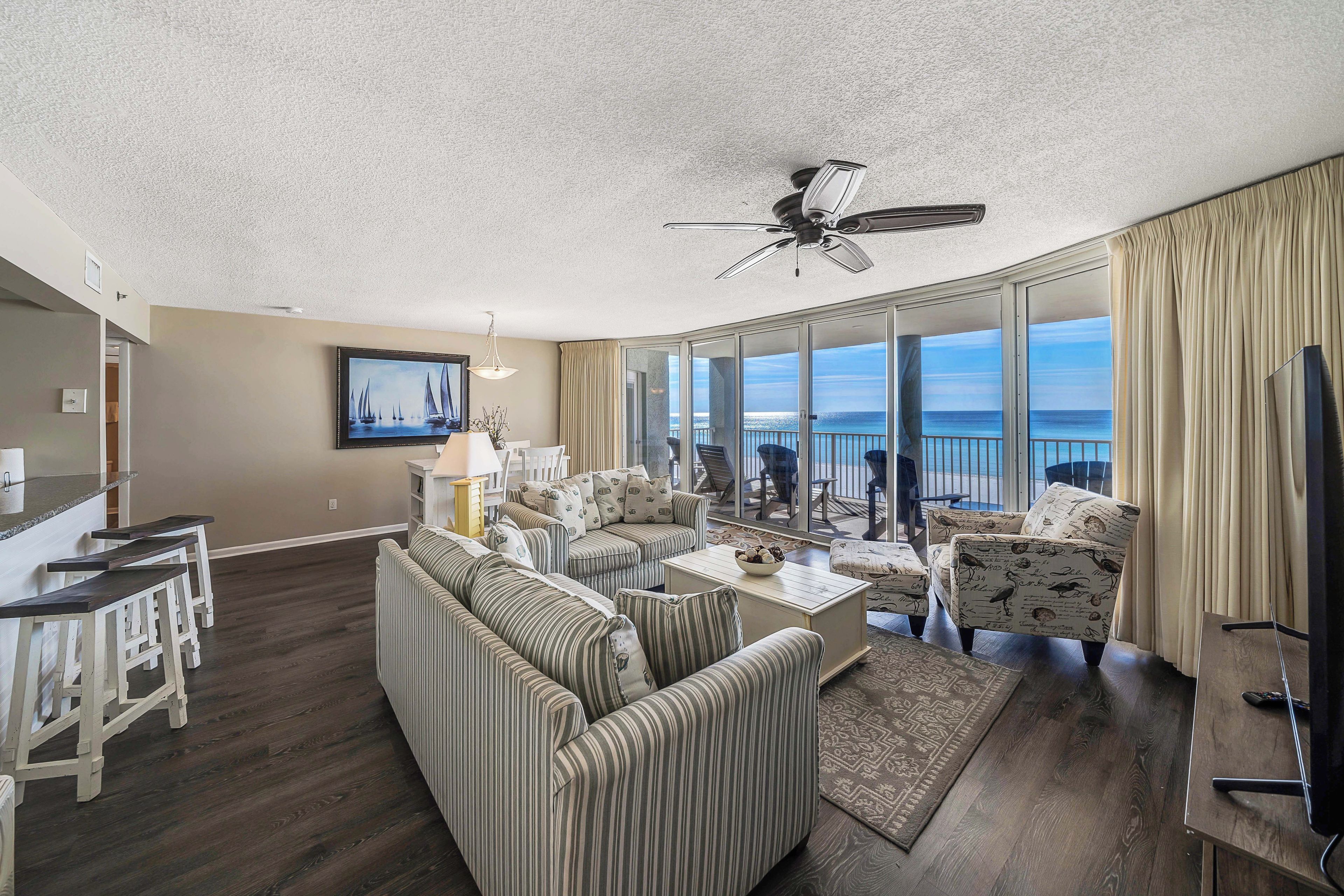 A spacious living area with a large window overlooking the ocean, a ceiling fan, and a dining area with bar stools.