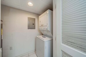 A laundry room with a front-loading washer and dryer, a small window with blinds, and a door with a handle.