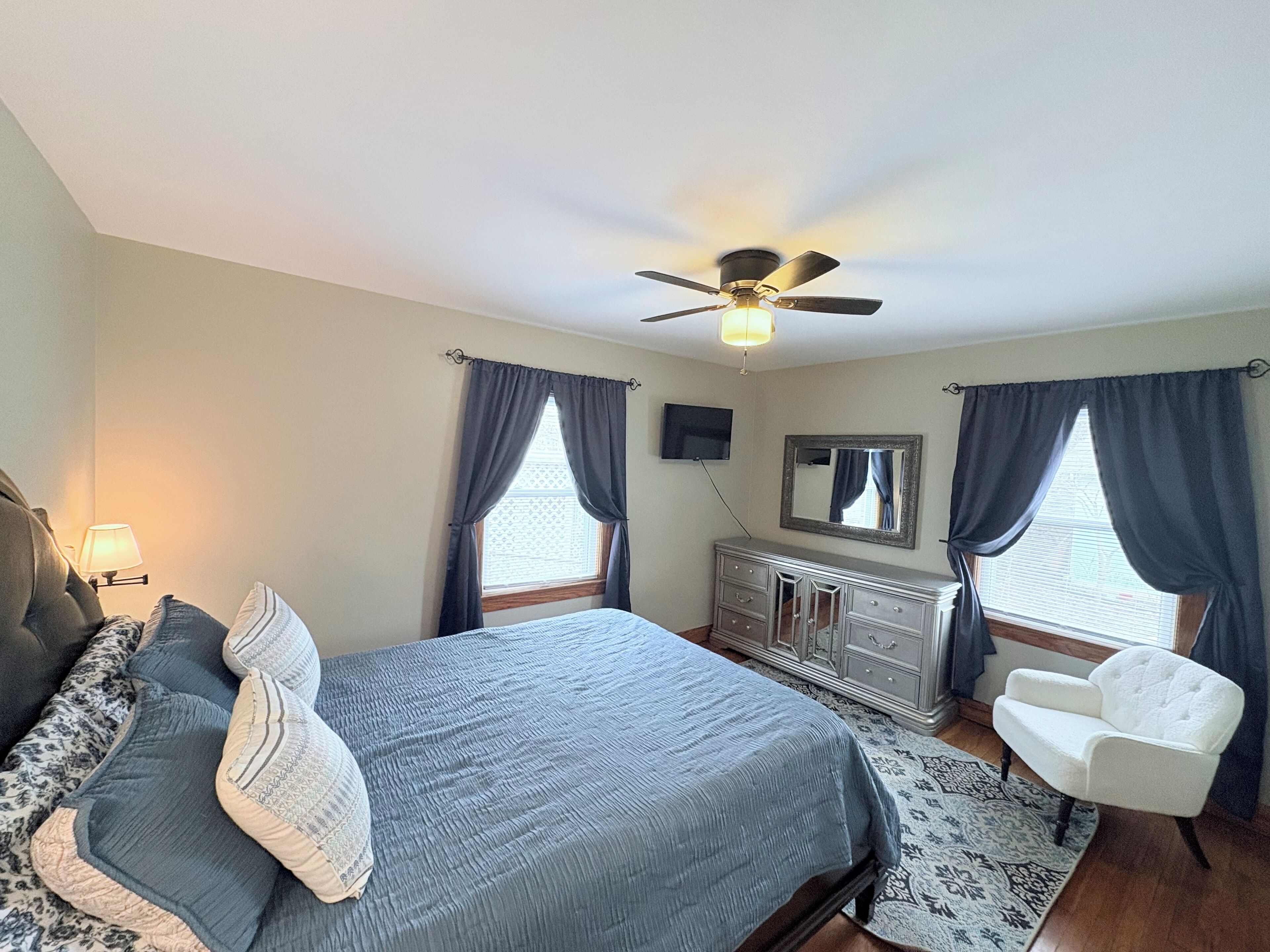 Une chambre à coucher comprenant un lit, une commode, une chaise, un ventilateur de plafond et un téléviseur.