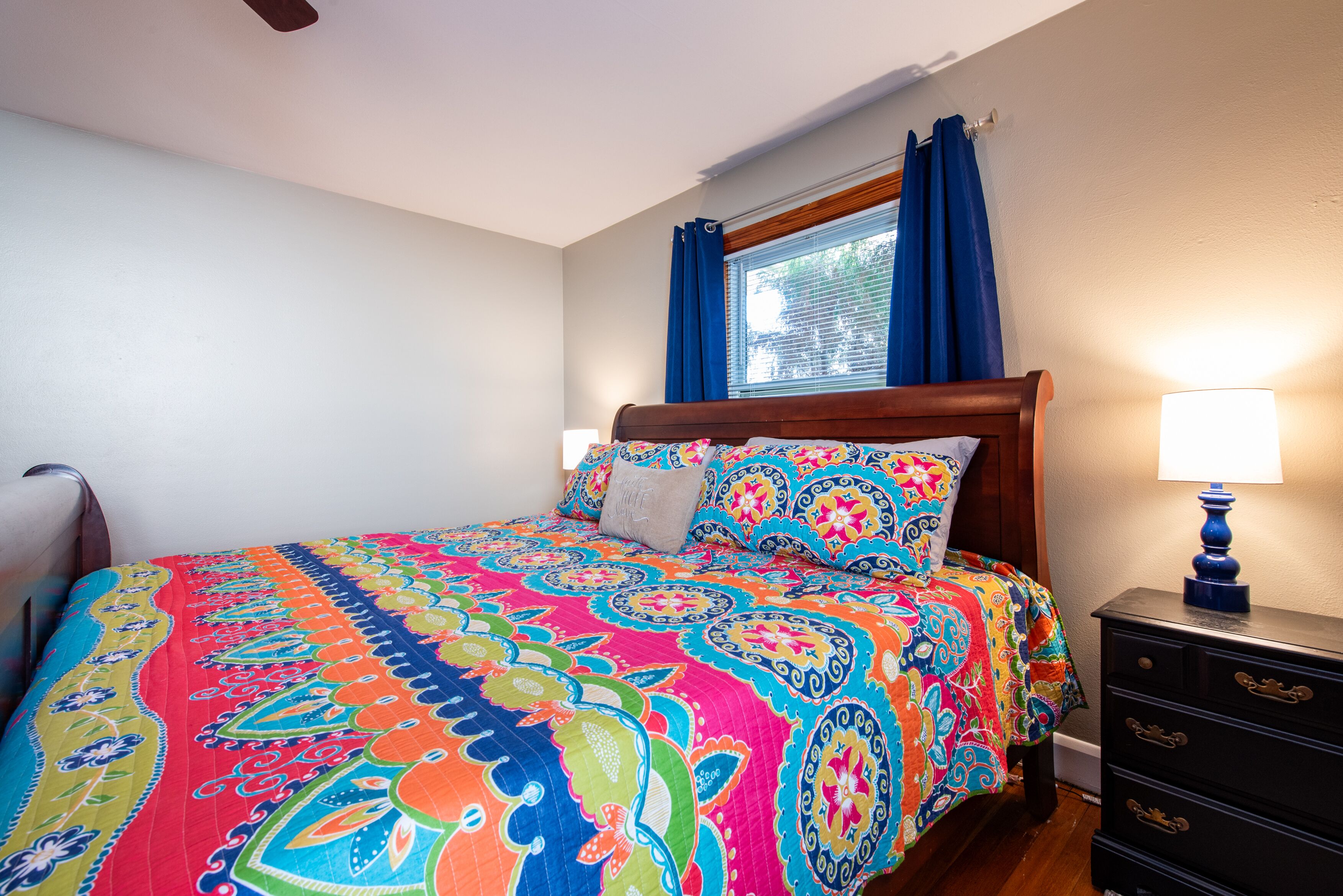 A bedroom with a colorful bedspread, a wooden headboard, a nightstand with a lamp, and a window with blue curtains.