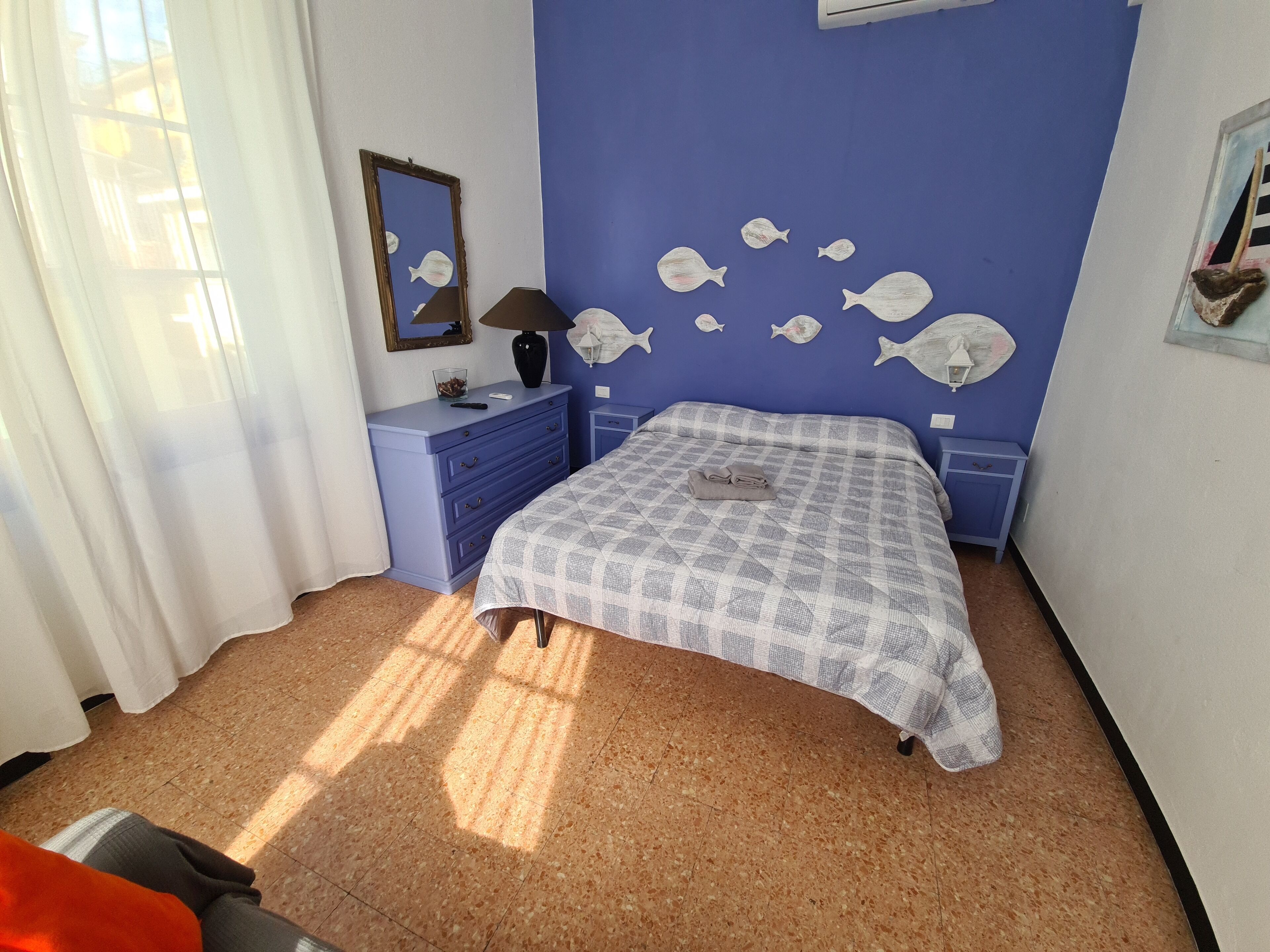 Un dormitorio con pared azul, cama con colcha a cuadros, cómoda, espejo y decoración de peces en la pared.