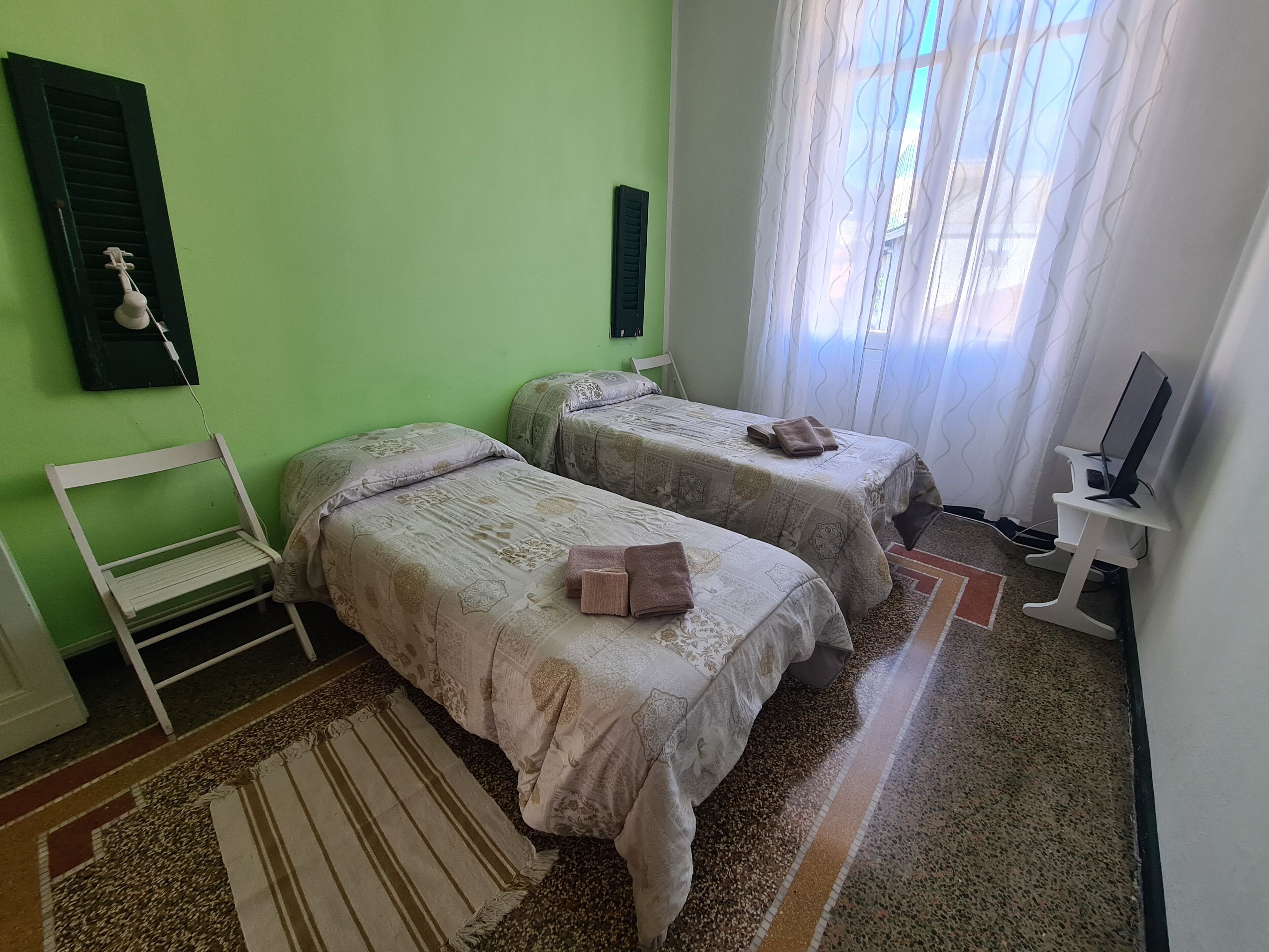 A room with two beds, a chair, a TV stand, and a window with curtains.
