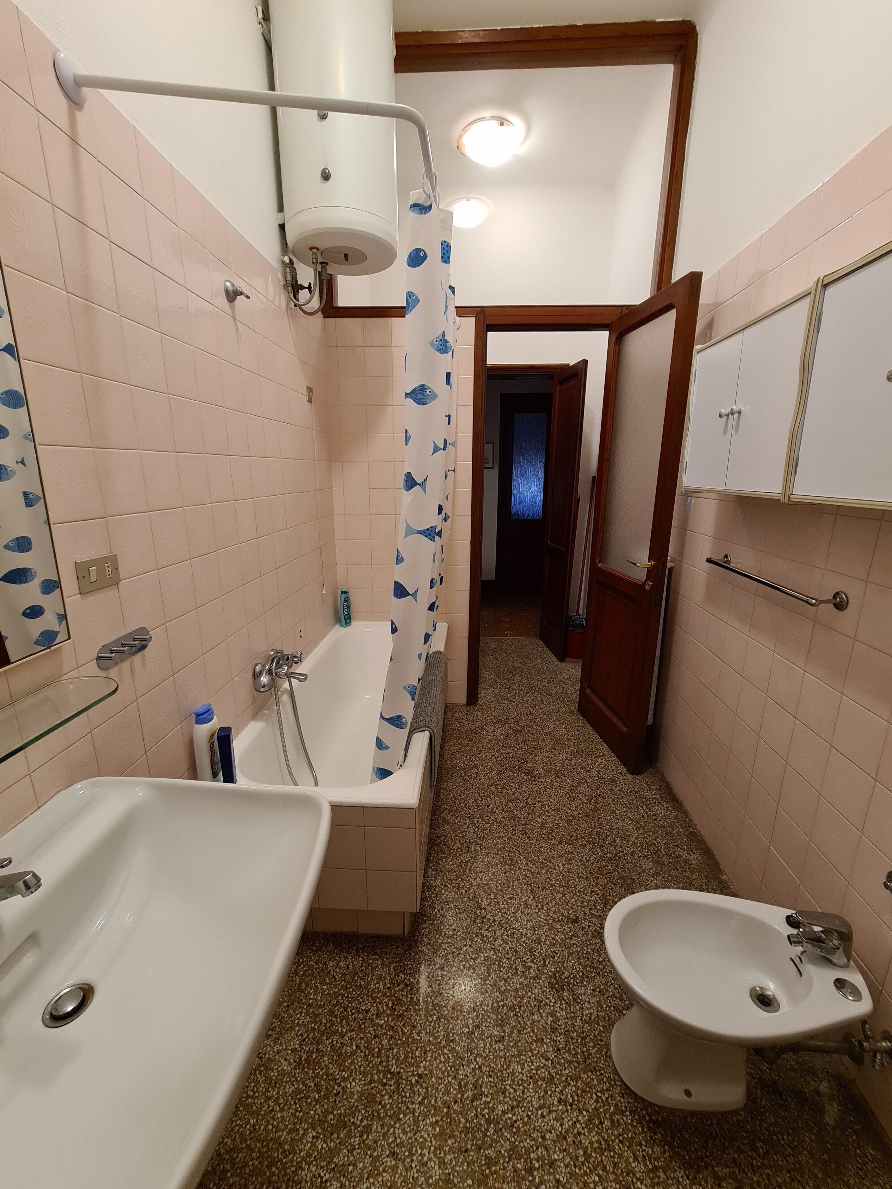 Un baño con bañera, lavamanos y bidé. La cortina de ducha tiene un estampado de peces y el piso está cubierto de baldosas con vetas.