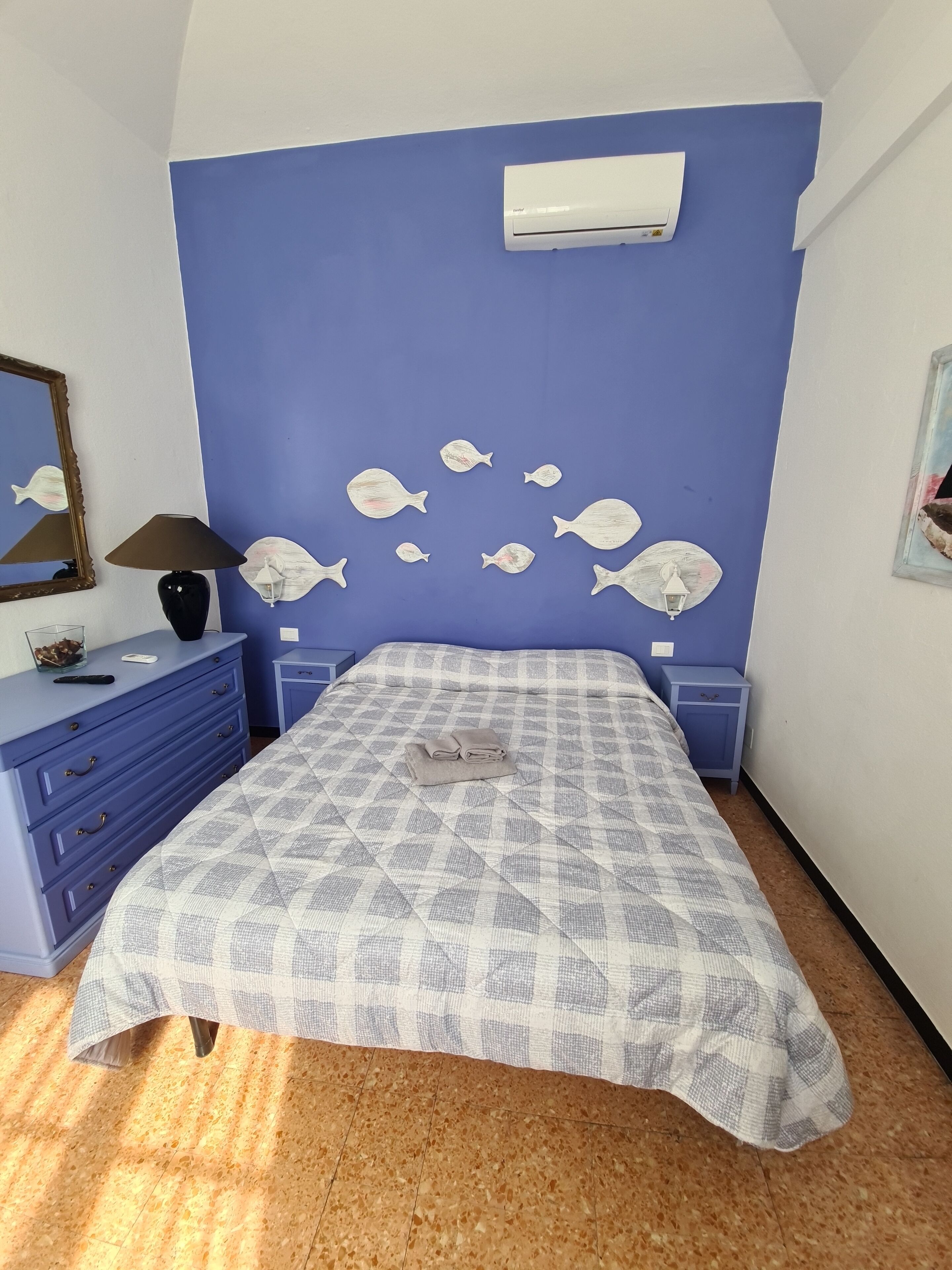 A bedroom with a blue wall, a bed with a checkered bedspread, a dresser, a nightstand, a lamp, a mirror, and wall art of fish.