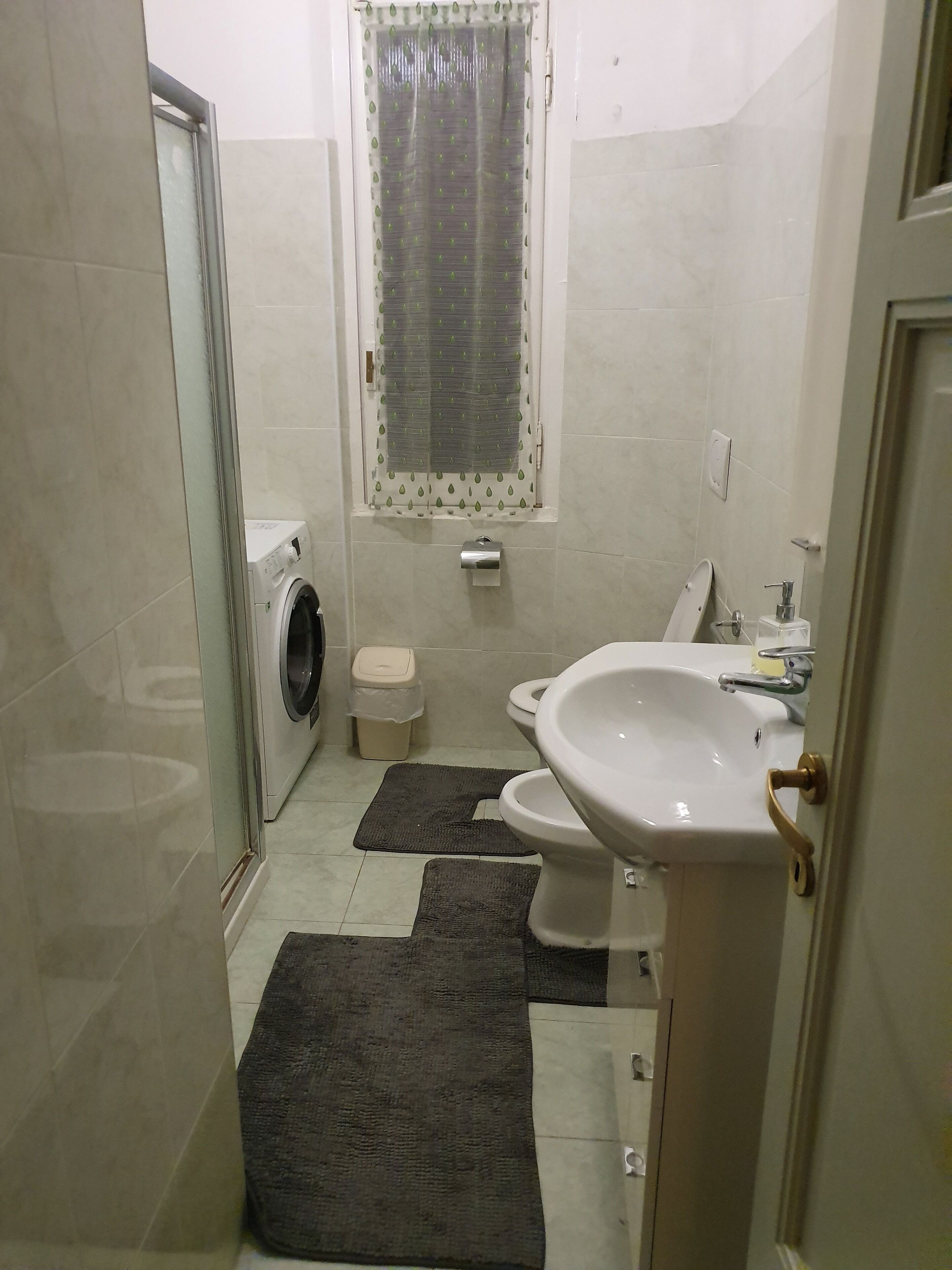 Badeværelse med vaskemaskine, toilet, håndvask og et vindue med gardin.