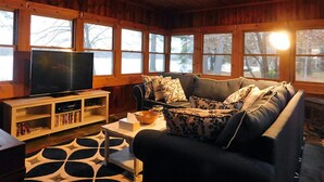 Ein gemütliches Wohnzimmer mit einem großen Flachbildfernseher, einem dunkelgrauen Sofa mit gemusterten Kissen und einer Holzbalkendecke.