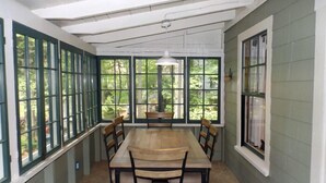 A dining area with a long table and chairs, large windows with green frames, and a hanging light fixture. - Charming Lakefront Cottage with Screened Porch & Shared Beach Access (Gray)