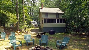 A cabin with a fire pit and Adirondack chairs in a wooded area. - Charming Lakefront Cottage with Screened Porch & Shared Beach Access (Gray)