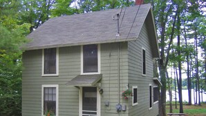 A two-story house with a front porch, a small garden, and a view of trees and a body of water. - Charming Lakefront Cottage with Screened Porch & Shared Beach Access (Gray)