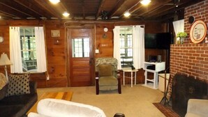 A cozy cabin interior with a wooden ceiling, a sofa, a chair, a small table, and a television. - Charming Lakefront Cottage with Screened Porch & Shared Beach Access (Gray)