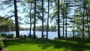 A lakeside view with a grassy area, a table, and chairs. - Charming cottage with stunning views and 180' of waterfront on Little Sebago. (Gray)