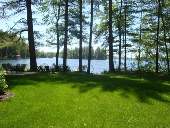 A lakeside view with a grassy area, a table, and chairs. - Charming cottage with stunning views and 180' of waterfront on Little Sebago. (Gray)