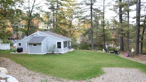 A small, single-story house with a covered porch and a well-maintained lawn. - Charming cottage with stunning views and 180' of waterfront on Little Sebago. (Gray)
