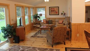 A cozy living room with a sofa, armchair, and coffee table, wooden floors, large windows, and a ceiling fan. - Charming cottage with stunning views and 180' of waterfront on Little Sebago. (Gray)