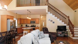 A spacious living area with a high ceiling, wooden staircase, and a kitchen with modern appliances. - Stunning modern home in peaceful, private setting on desirable Long Lake. (Bridgton)