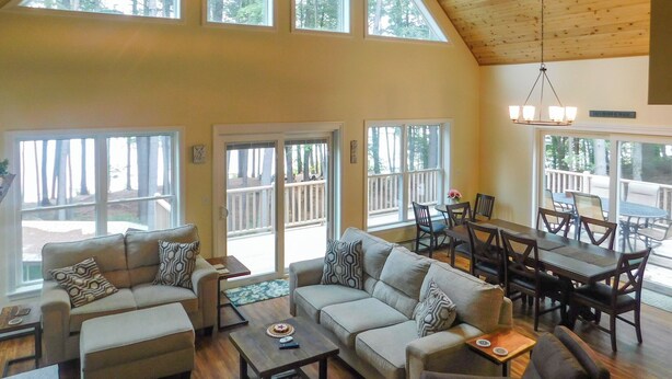 A spacious living area with a high ceiling, large windows, and a dining area with wooden furniture. - Stunning modern home in peaceful, private setting on desirable Long Lake. (Bridgton)