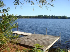 Un muelle de madera que se adentra en un lago tranquilo, rodeado de árboles y una costa boscosa a lo lejos.