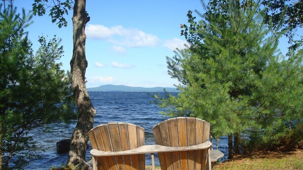 Zwei Holz-Adirondack-Stühle, die auf ein Gewässer mit Bäumen und eine ferne Landschaft zeigen.