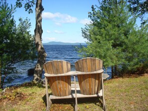 To træstole af Adirondack-typen står med ryggen mod et vandareal, med træer og et fjerntliggende landskab i baggrunden.
