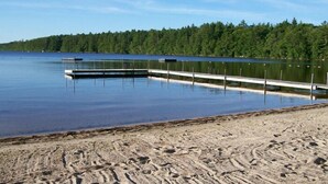 Une plage de sable bordée d’un lac paisible, avec un quai et une forêt en arrière-plan.