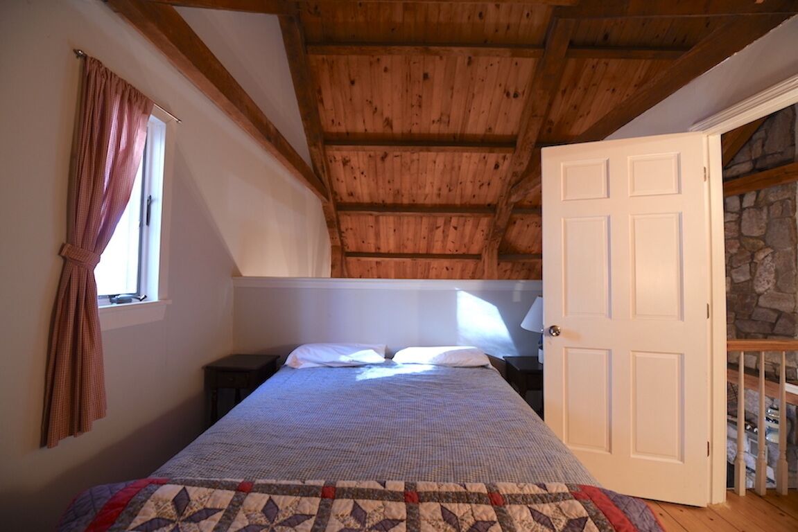 Quarto com teto de madeira, cama com colcha, mesa de cabeceira, janela com cortinas e parede de pedra.