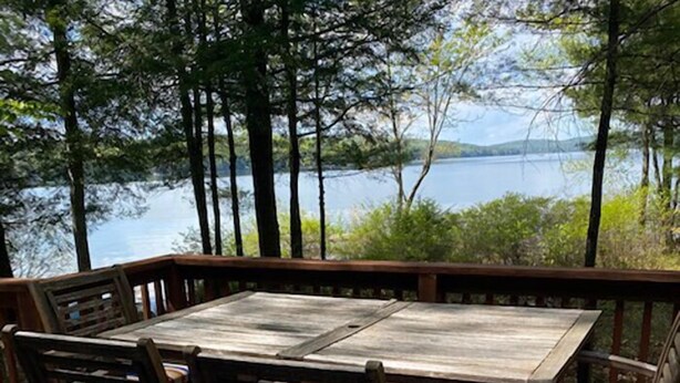 Une terrasse en bois avec une table et des chaises, donnant sur un lac, avec des arbres et des collines en arrière-plan.