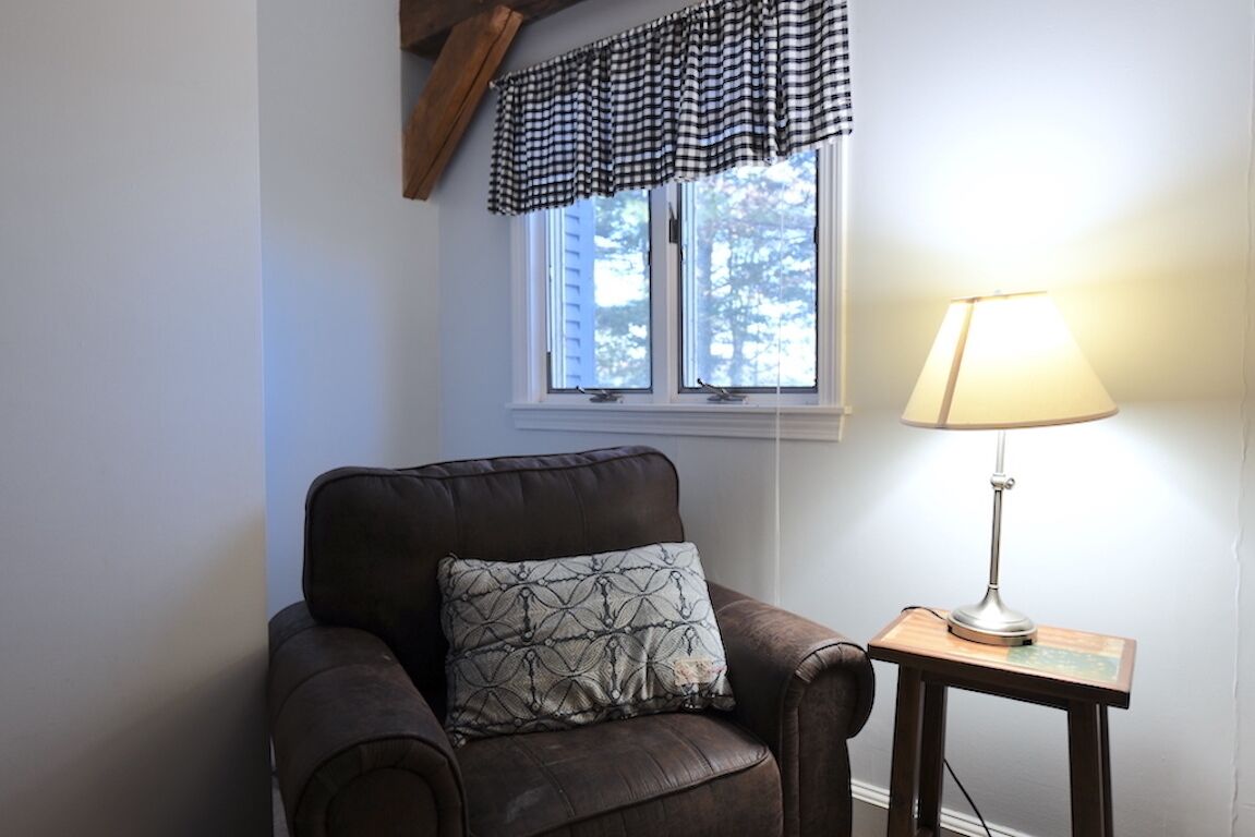 A room with a brown leather armchair, a patterned pillow, a window with a checkered valance, and a table lamp.