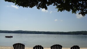 Three Adirondack chairs on a sandy beach with a calm lake and a boat in the distance.