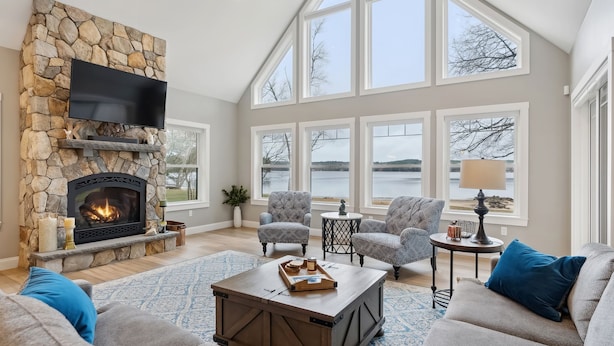 A spacious living room with a stone fireplace, comfortable seating, large windows, and a view of a lake.