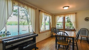 A room with a piano, dining table, chairs, and large windows overlooking a body of water. - Spacious, cozy waterfront cottage on Norway Lake with hiking trails nearby. (Norway)