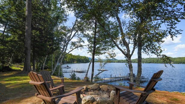 Two Adirondack chairs facing a lake with a dock and dock chairs, surrounded by trees and a clear sky. - Spacious, cozy waterfront cottage on Norway Lake with hiking trails nearby. (Norway)