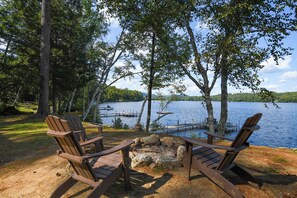 Deux fauteuils Adirondack sont placés face à un lac, avec une jetée et des chaises de jetée, le tout entouré d’arbres et sous un ciel dégagé.