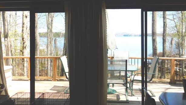 A view from a room with large windows overlooking a lake, with outdoor furniture and a wooden deck.